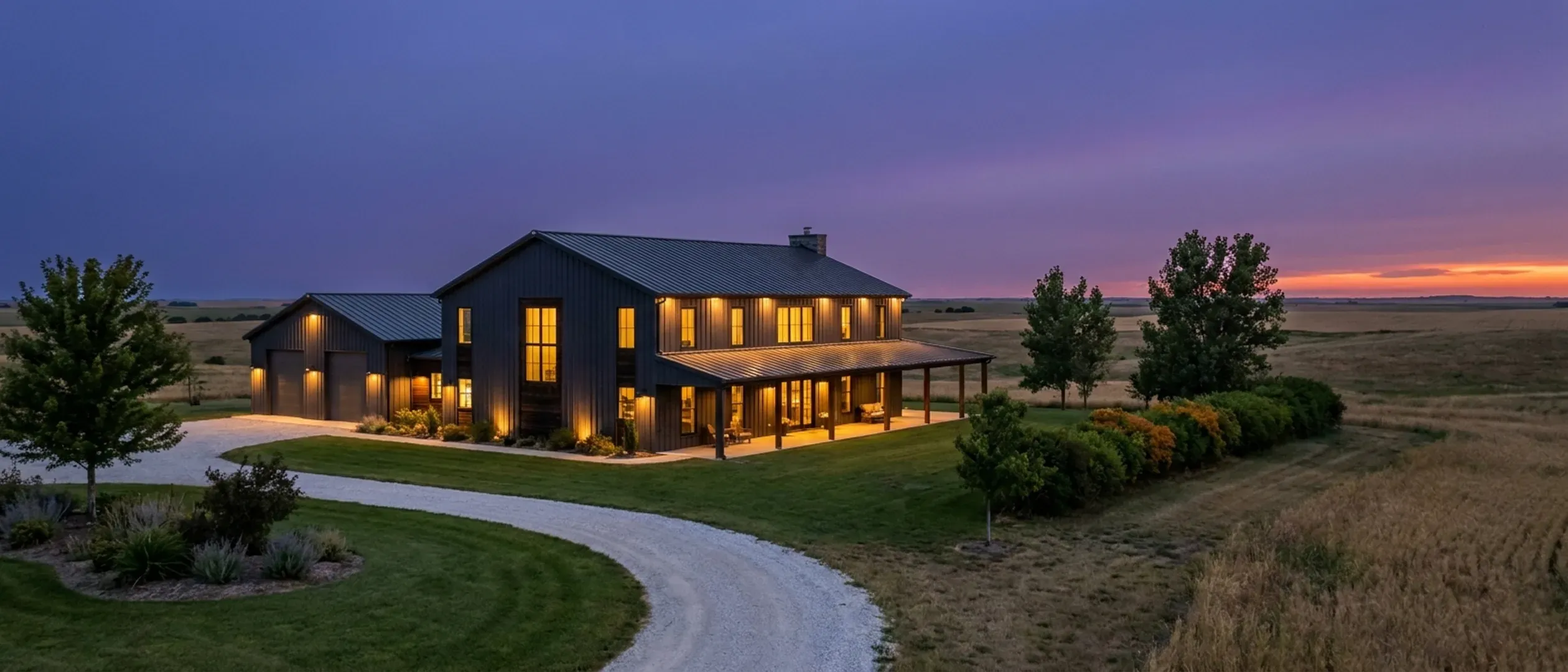 A barndominium at dusk with warm light glowing from every window and exterior lighting along the porch and roofline against a deep blue twilight sky.